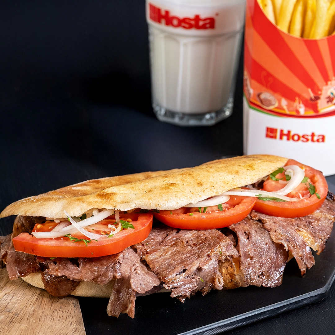 Zemin üzerinde özenle yerleştirilmiş ekmek arası et dönerin arka planında bulanık şekilde patates kızartması ve bir bardak ayran görülüyor.