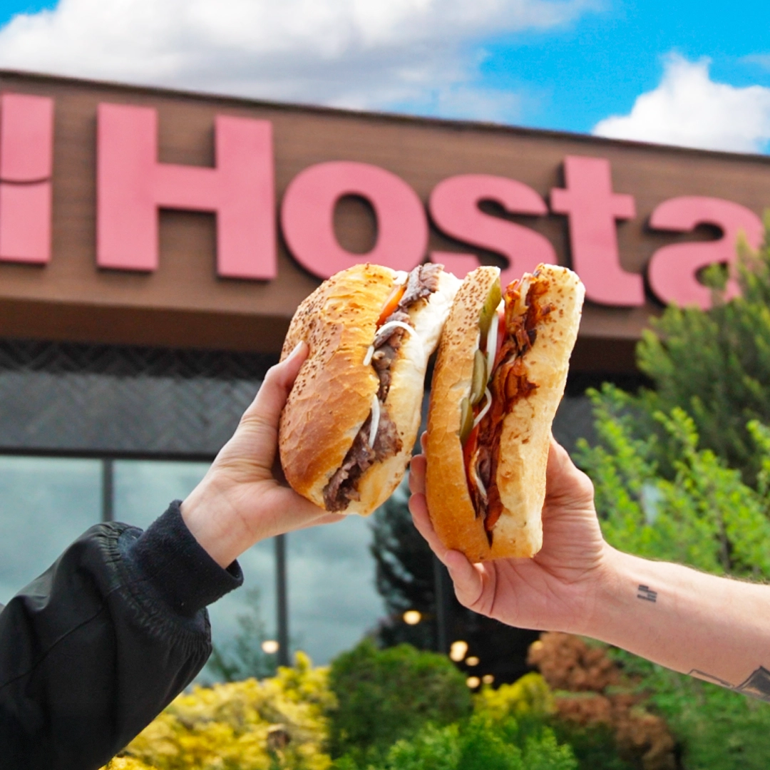 İki kişi ellerindeki ekmek arası dönerleri tutarken arka planda bulanık şekilde bir Hosta Döner şubesi görülüyor.