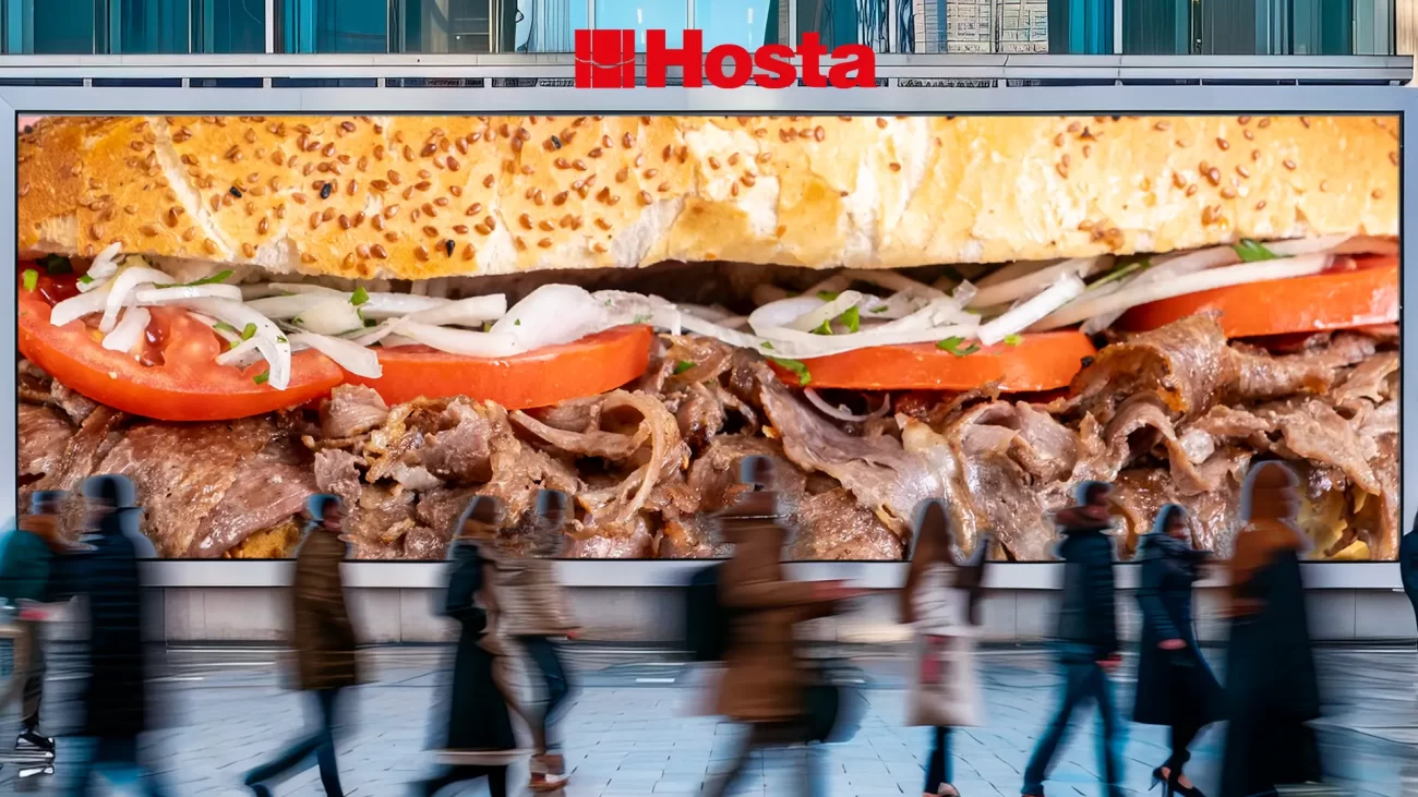 Sokakta, arka planda Hosta ekmek arası et döner görseli bulunan billboardın önünden geçen insanlar.